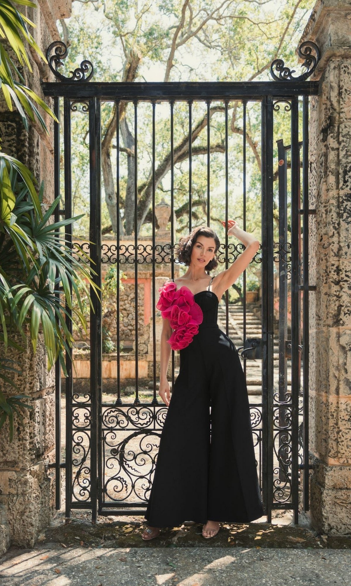 A woman wearing the Long Formal Jumpsuit 11767 by Mac Duggal, featuring a black wide-leg silhouette with pink floral detail, stands before an ornate wrought-iron gate and stone pillars amid lush greenery.